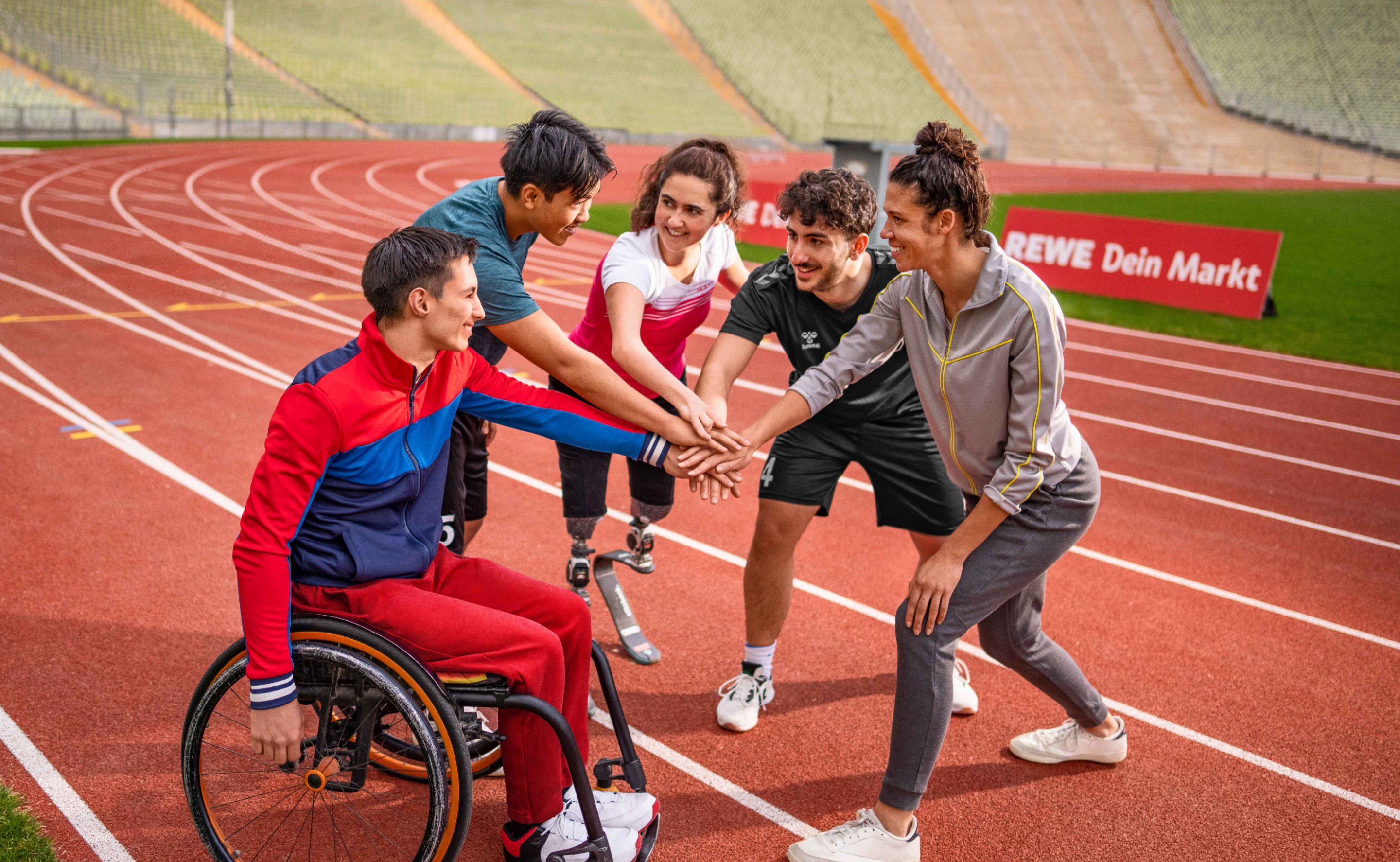 Menschen mit und ohne Behinderung legen ihre Hände aufeinander, um sich auf einer Tartanbahn auf ein sportliches Event einzustimmen.
