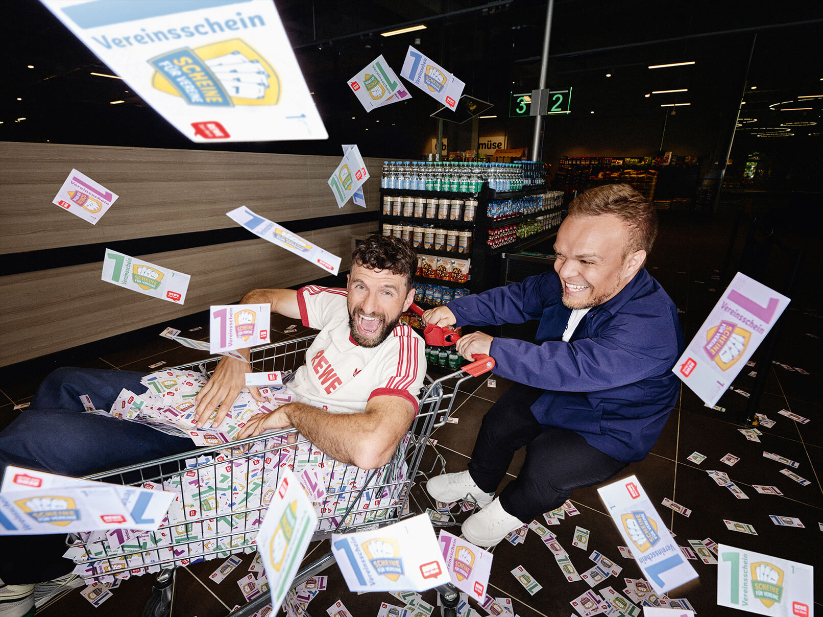 Thomas Müller sitzt in einem Einkaufswagen voller Vereinsscheine und wird von Niko Kappel durch einen REWE Markt geschoben. Die beiden haben Spaß und Vereinsscheine fliegen durch die Luft.