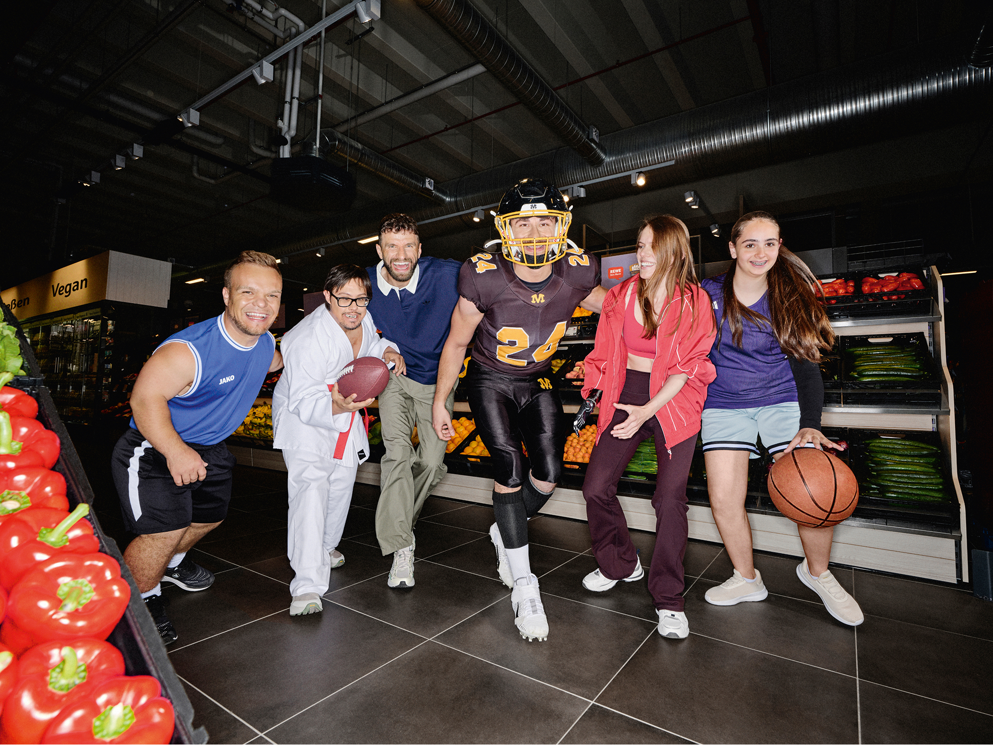 In einem REWE Markt haben Niko Kappel und Thomas Müller gemeinsam Spaß beim Posieren mit diversen Sportler:innen in ihren Sportoutfits.