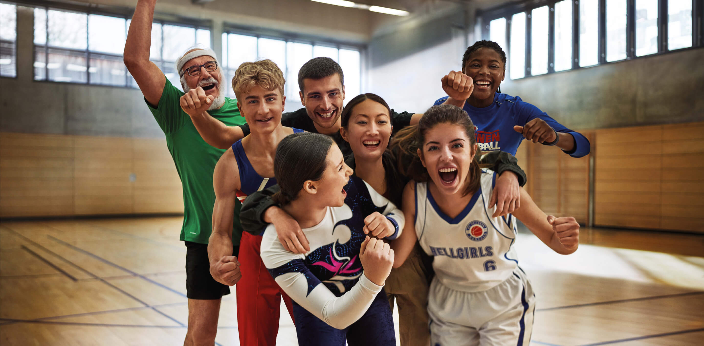 Sieben diverse Sportler:innen stehen jubelnd in einer Turnhalle.