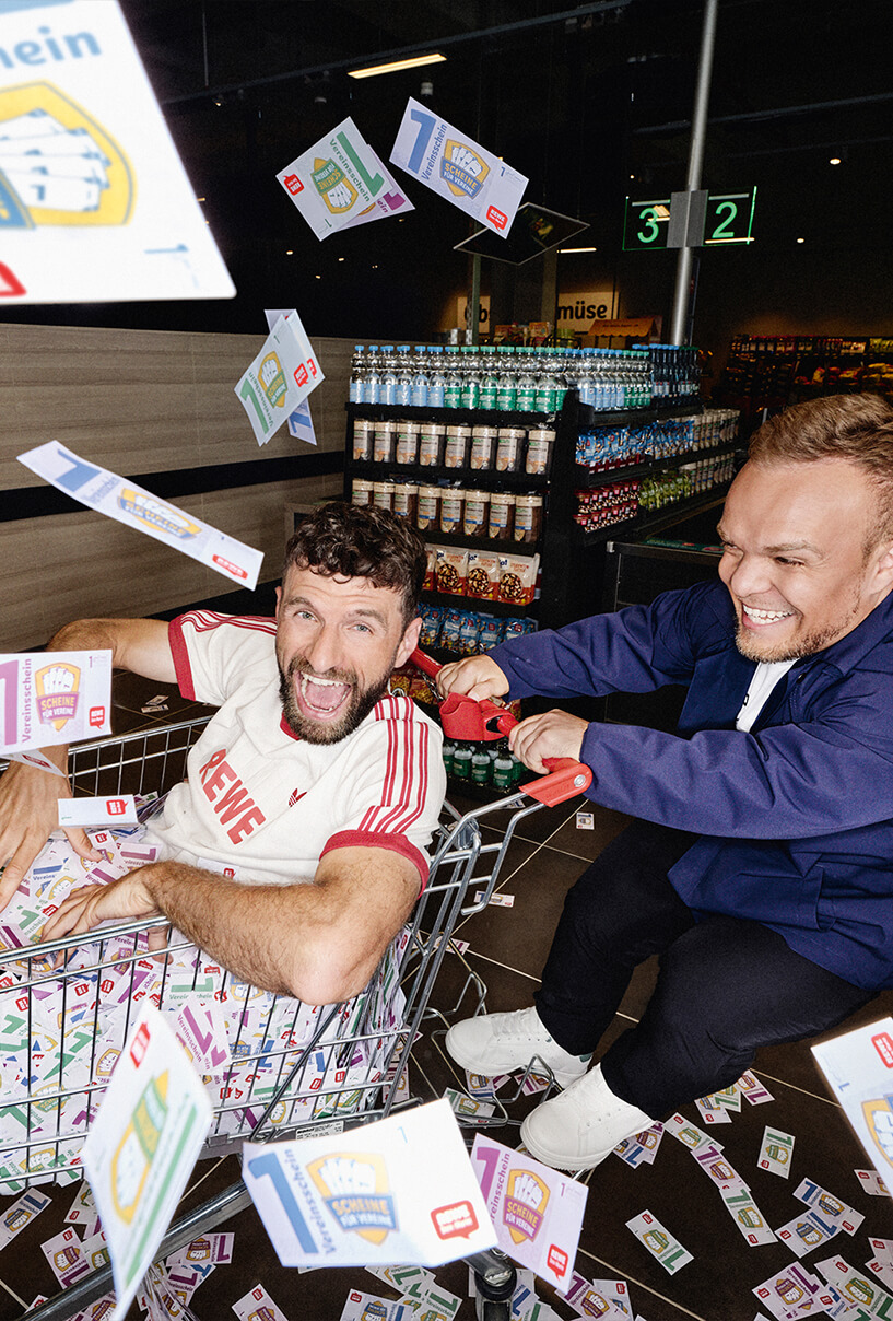 Thomas Müller sitzt in einem Einkaufswagen voller Vereinsscheine und wird von Niko Kappel durch einen REWE Markt geschoben. Die beiden haben Spaß und Vereinsscheine fliegen durch die Luft.