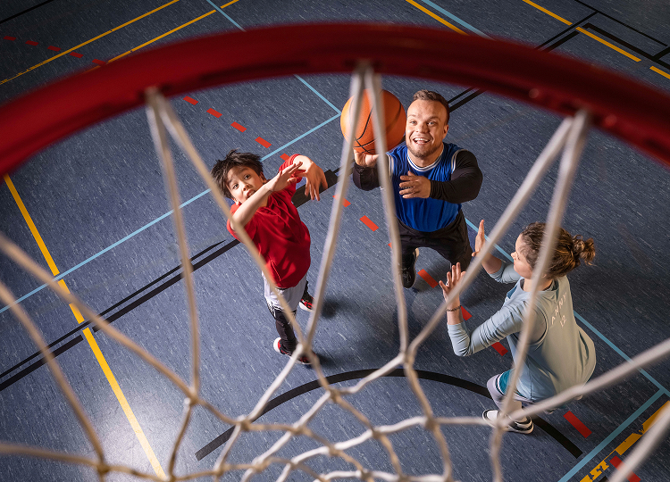Para-Sportler Niko Kappel sowie ein Mädchen und ein Junge spielen gemeinsam in einer Turnhalle Basketball. Mit Blickrichtung von oben durch den Basketball-Korb ist der Moment zu sehen  sehen, als Niko auf den Korb wirft.