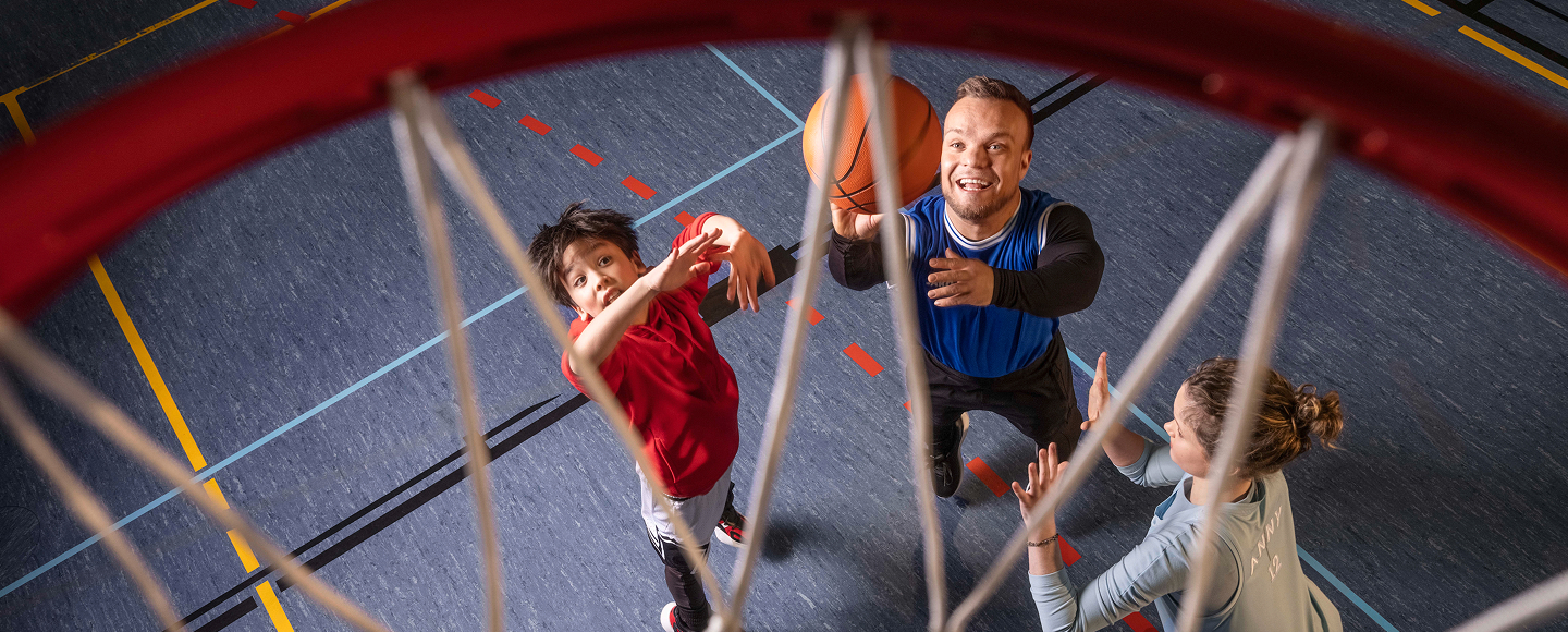 Para-Sportler Niko Kappel sowie ein Mädchen und ein Junge spielen gemeinsam in einer Turnhalle Basketball. Mit Blickrichtung von oben durch den Basketball-Korb ist der Moment zu sehen  sehen, als Niko auf den Korb wirft.