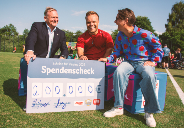 REWE übergibt Teil-Spende an die Aktion Mensch: Peter Maly (REWE Group), Para-Weltmeister Niko Kappel und Christina Marx (Aktion Mensch) sitzen lachend auf einem Rasen-Sportplatz und halten einen großen Spendenscheck über 200.000 €