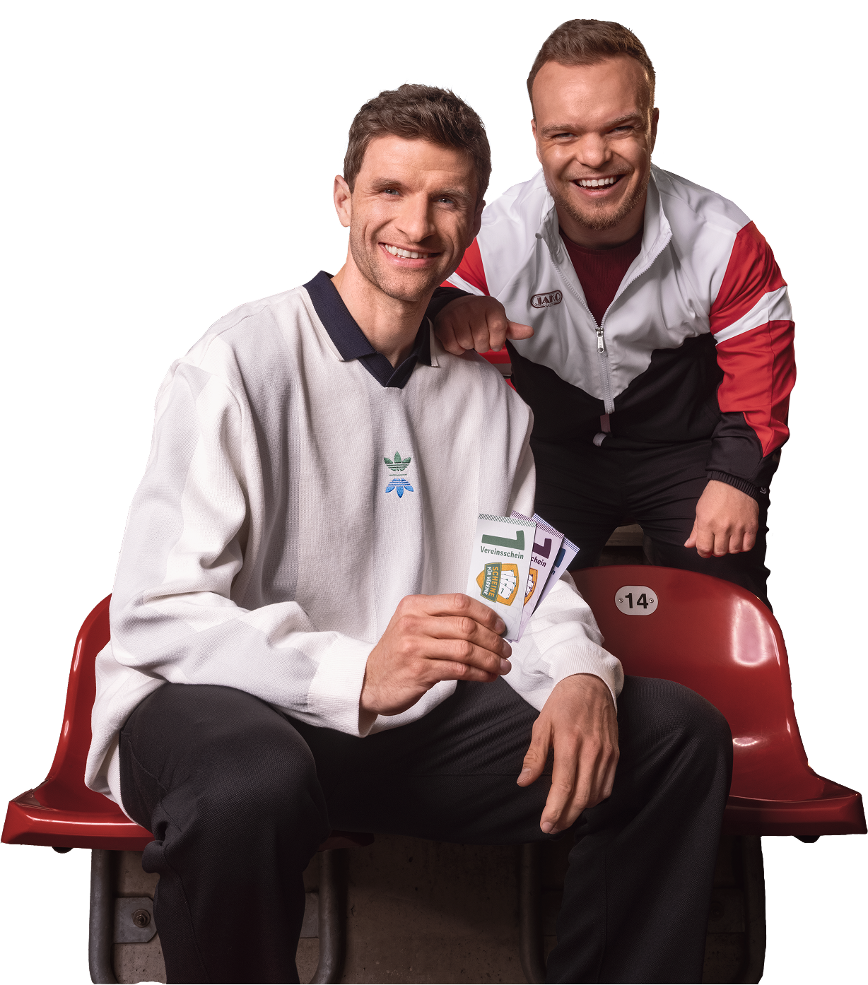 Thomas Müller und Niko Kappel sitzen lachend auf einer Stadiontribüne und zeigen ihre Vereinsscheine.